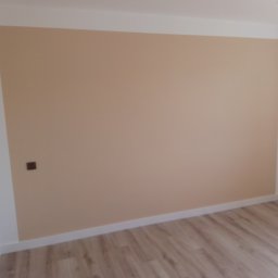 Bizoń Rafał usługi remontowo budowlane - Ściana pomalowana na beżowo z brązowym włącznikiem światła i fragmentem podłogi wyłożonej panelami w odcieniach brązu.