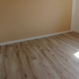 Jasne panele podłogowe imitujące drewno, świeżo położone w pokoju z beżowymi ścianami i białymi listwami przypodłogowymi, widoczny brązowy włącznik elektryczny.