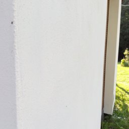 Bizoń Rafał usługi remontowo budowlane - Fragment ściany budynku z jasną, chropowatą fakturą, widoczny narożnik i fragment okna z brązową ramą, w tle trawa i zieleń ogrodu.