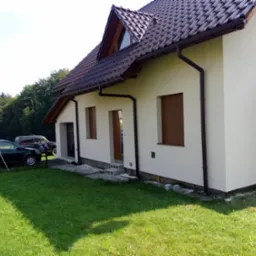 Jasny dom jednorodzinny z brązową dachówką i rynnami, widok z boku na trawnik i dwa samochody w tle.