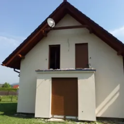 Otynkowana ściana szczytowa domu z brązową stolarką okienną i satelitą na dachu, widoczne ślady świeżego tynku przy gruncie.