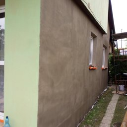 Bizoń Rafał usługi remontowo budowlane - Ściana budynku po nałożeniu nowej warstwy szpachli, z widocznym fragmentem ocieplenia w kolorze pistacjowym, zabezpieczone okna, rusztowanie w tle i butelka wody mineralnej przy ścianie.