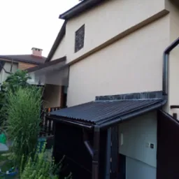 Elewacja domu z widoczną zabudową nad wejściem, rynny spustowe i fragment ogrodu z roślinnością