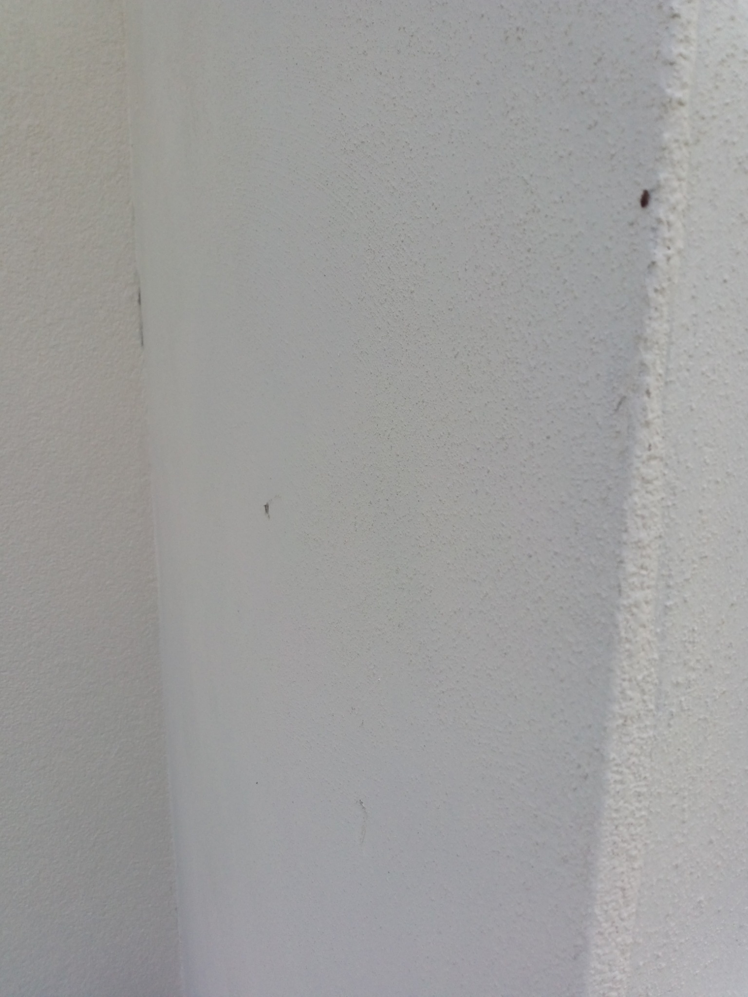 Biała ściana w rogu pomieszczenia z widoczną fakturą i drobnymi niedoskonałościami, punktowe zabrudzenia na powierzchni.