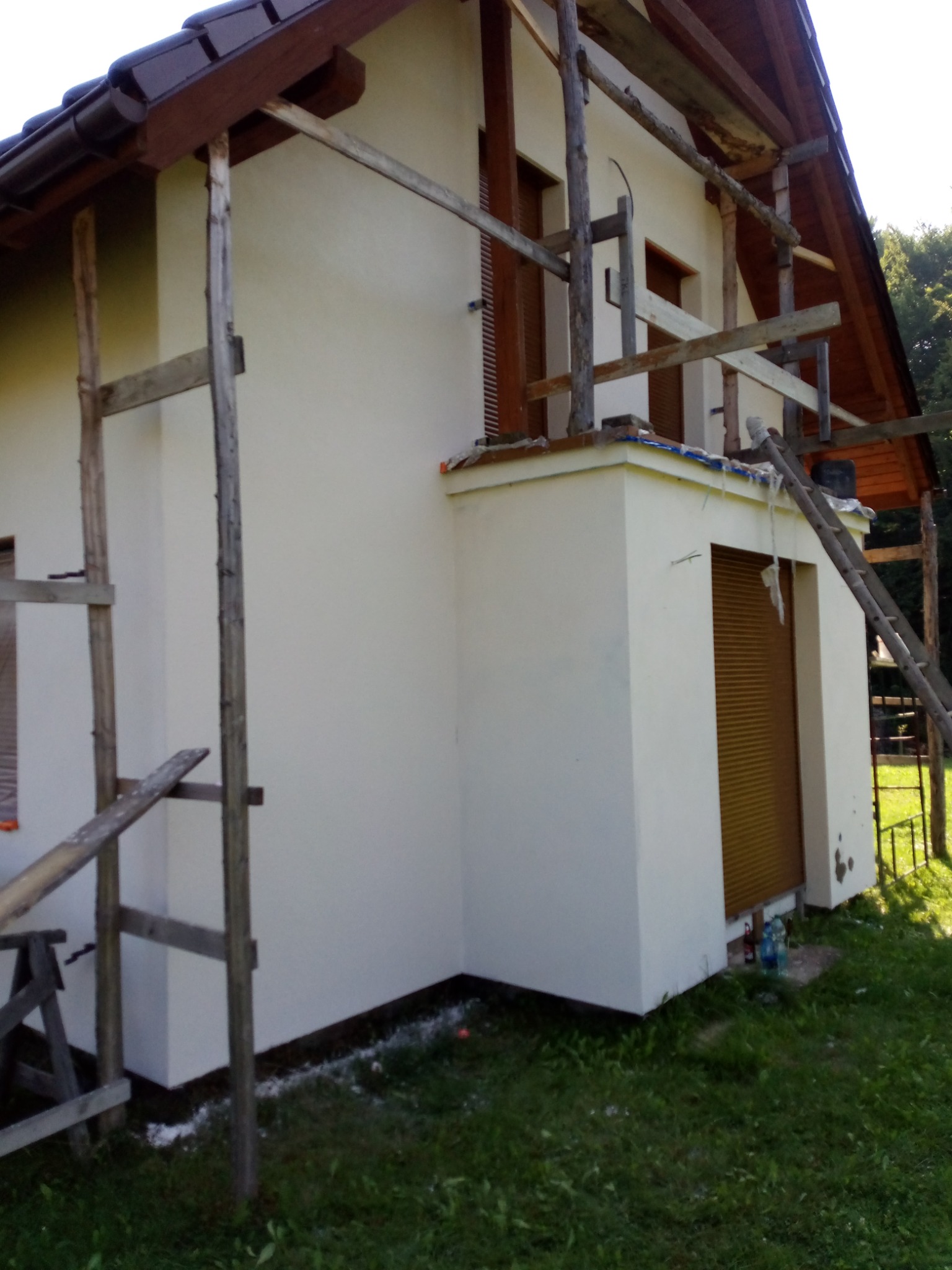 Dom jednorodzinny w trakcie prac wykończeniowych elewacji, widoczne drewniane rusztowania, świeżo nałożona jasna warstwa tynku, zabezpieczone okna roletami zewnętrznymi.