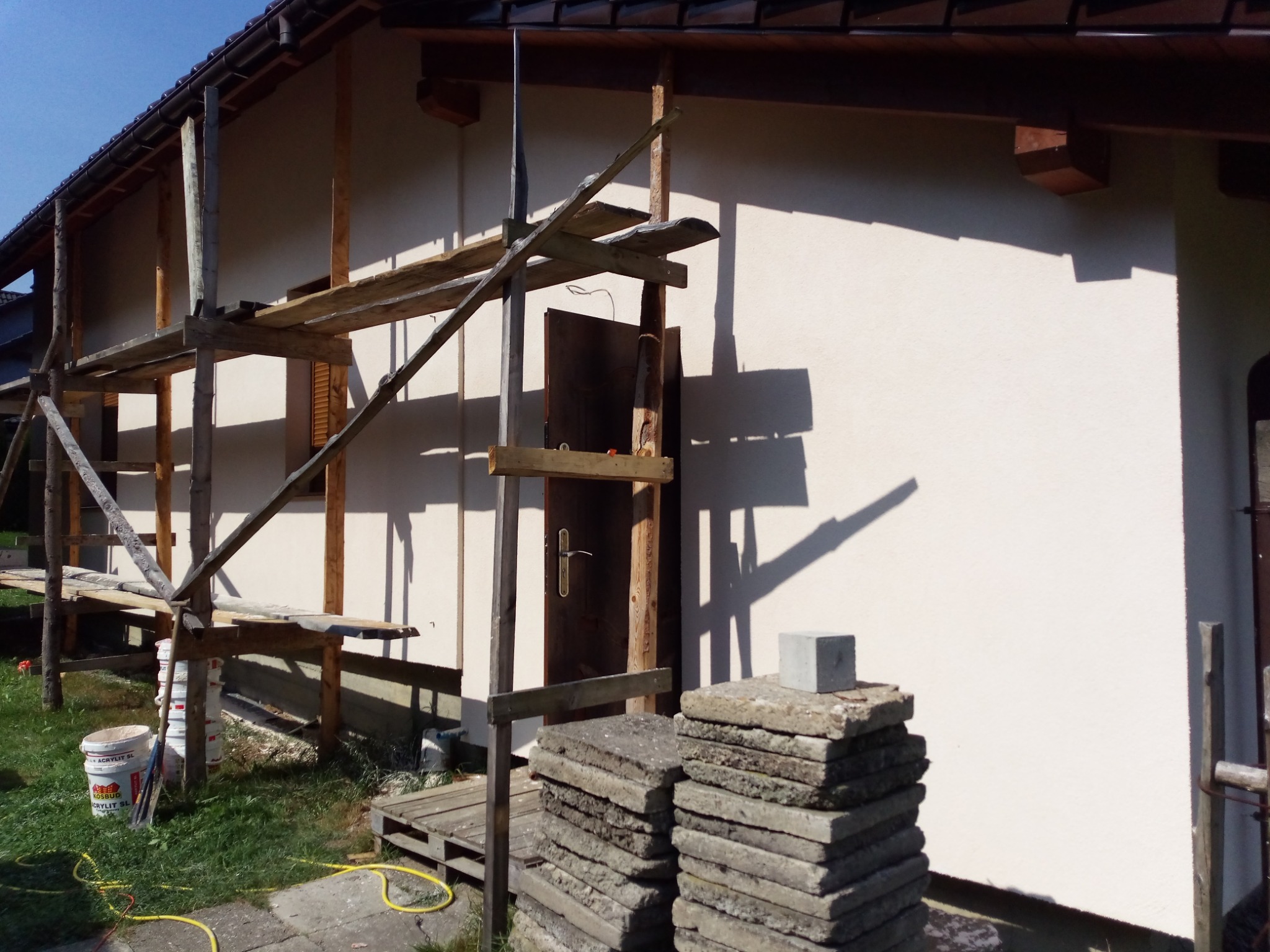 Elewacja domu w trakcie prac wykończeniowych, widoczne drewniane rusztowanie, sterta płyt chodnikowych i wiadro z farbą akrylową.