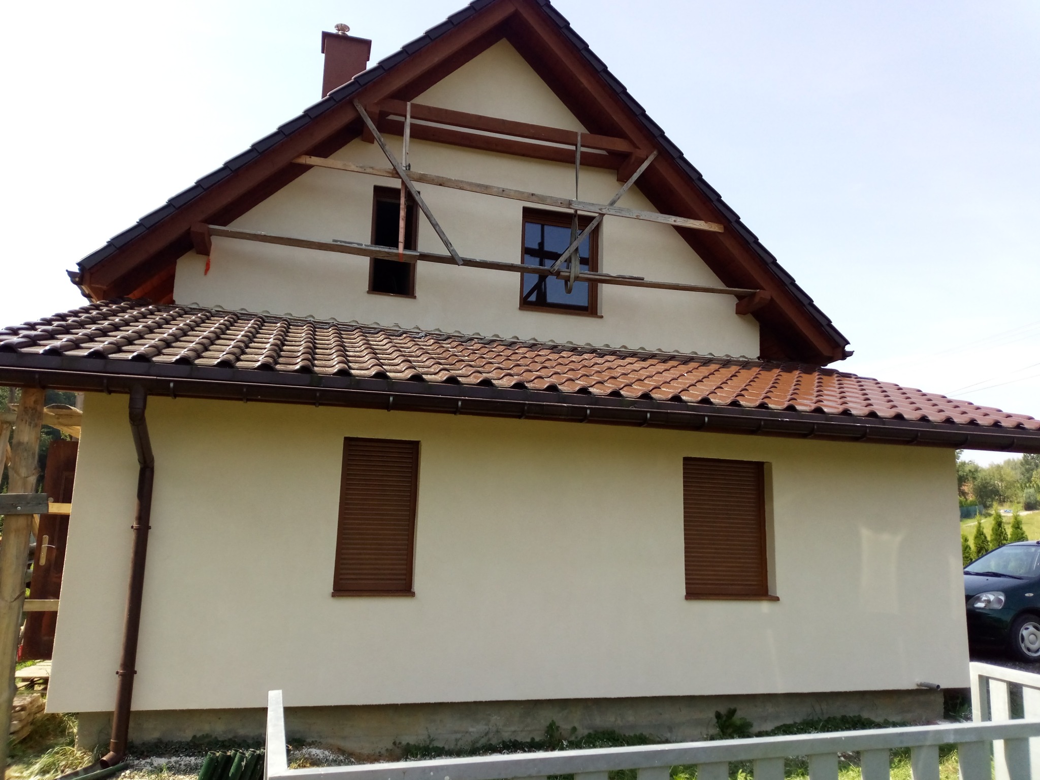 Dwupiętrowy dom z jasną elewacją, brązową dachówką i roletami w oknach, z widocznym rusztowaniem na poddaszu i częściowo odsłoniętym fundamentem.
