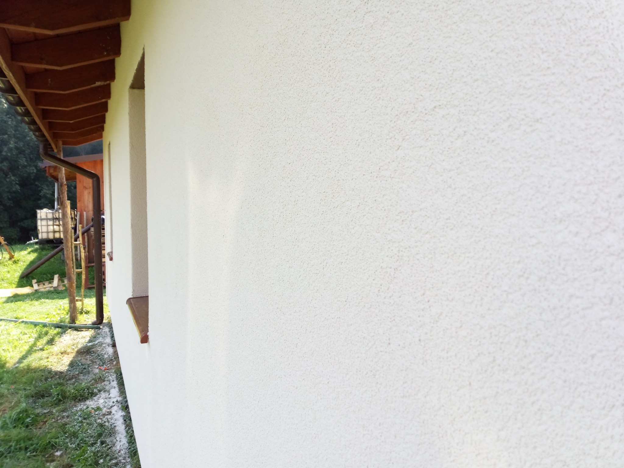 Ściana budynku z jasnym, ziarnistym tynkiem zewnętrznym, widoczny fragment dachu z brązowymi elementami drewnianymi, okno z brązowym parapetem i rynna w tle, trawa i zielona roślinność u podstawy...