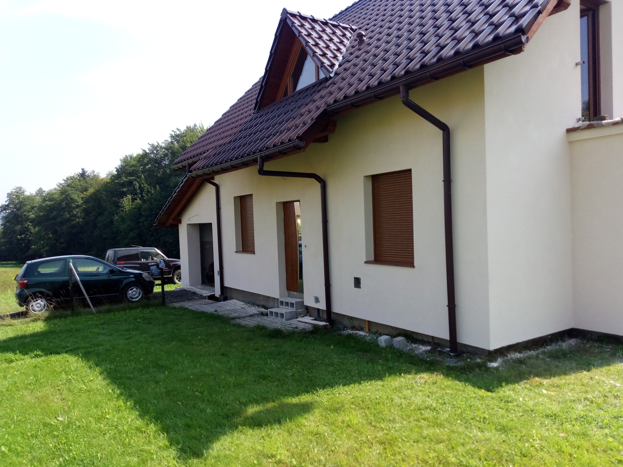 Jasny dom jednorodzinny z brązową dachówką i rynnami, widok z boku na trawnik i dwa samochody w tle.