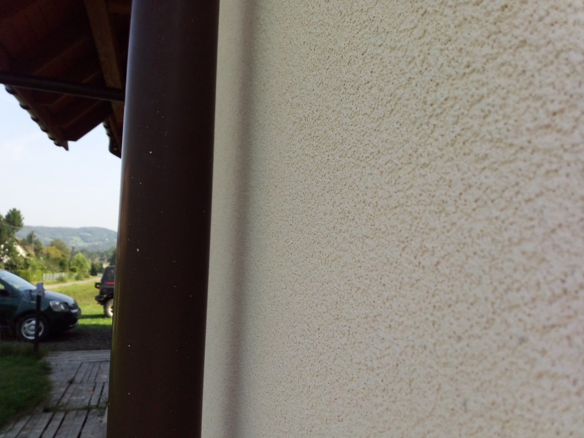 Beżowa ściana zewnętrzna z fakturą baranka, brązowa rynna pionowa, w tle zielony krajobraz z samochodami na podjeździe.