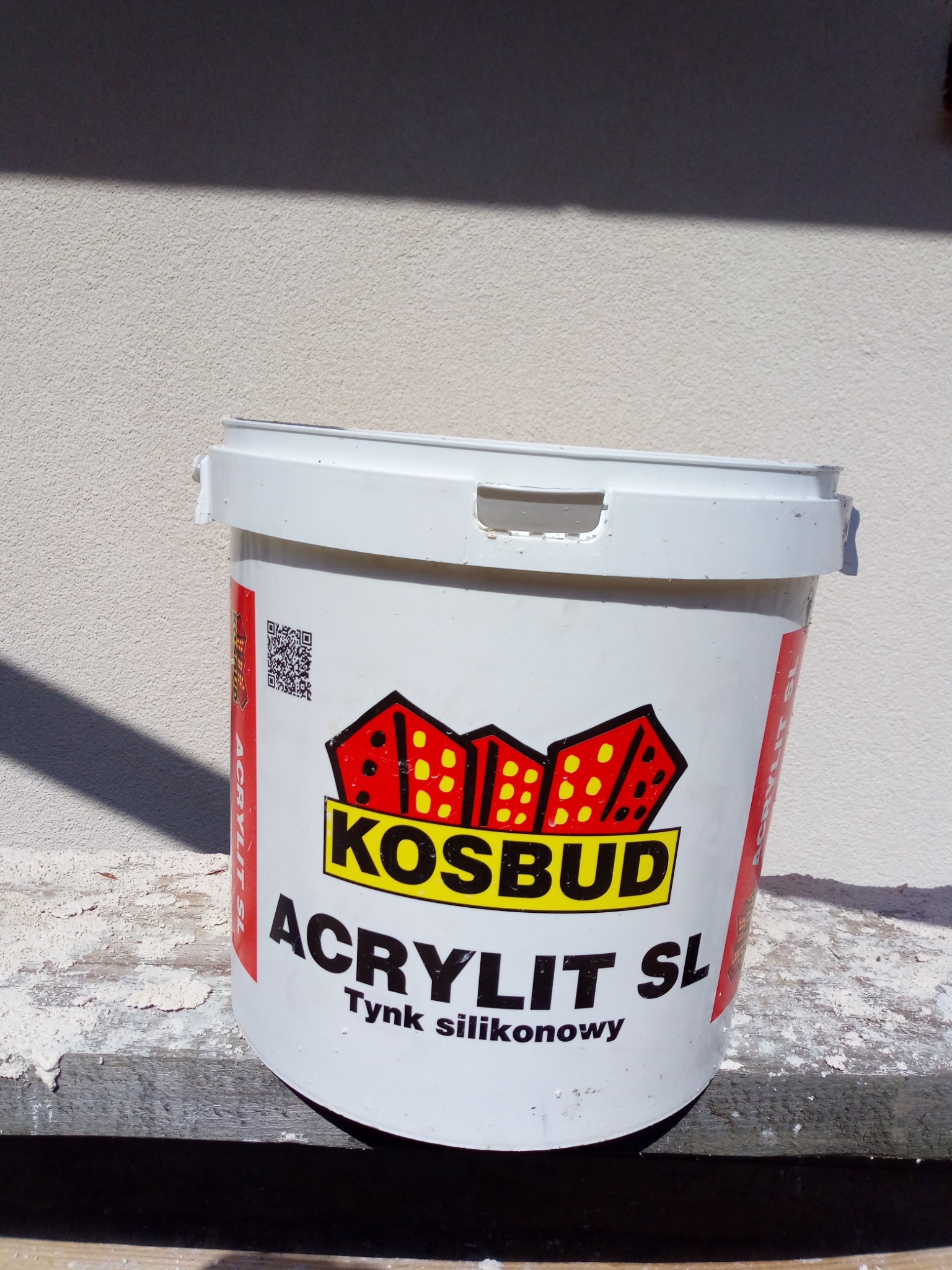 Wiadro z silikonowym tynkiem 'Acrylit SL' firmy Kosbud, stojące na drewnianej powierzchni, z widoczną strukturą tynku na ścianie w tle.