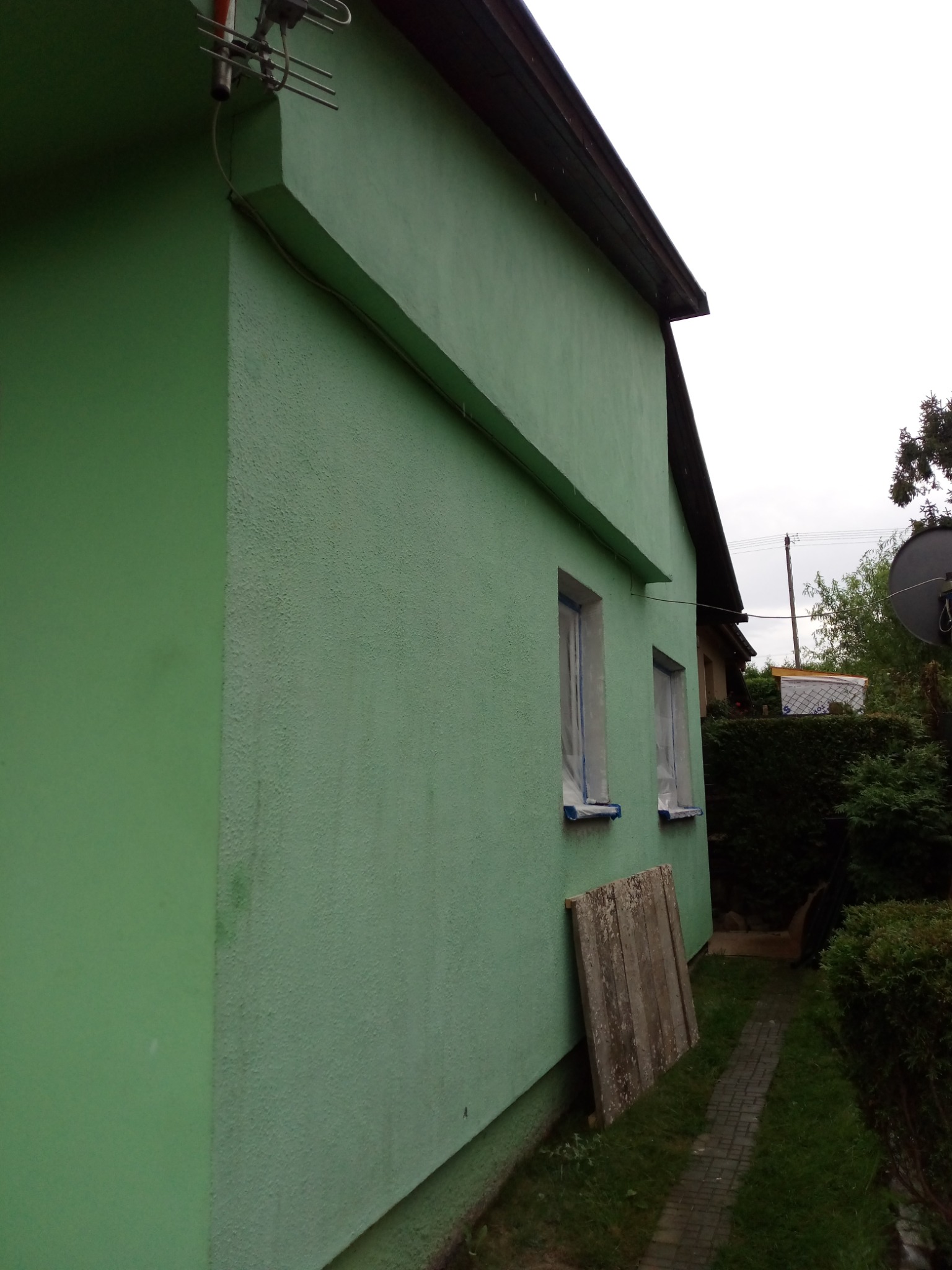 Ściana domku działkowego po malowaniu na zielono, z zabezpieczonymi oknami folią malarską. Fragment ściany ma inną fakturę i odcień zieleni. Przy ścianie oparte płyty. Widoczny fragment dachu...