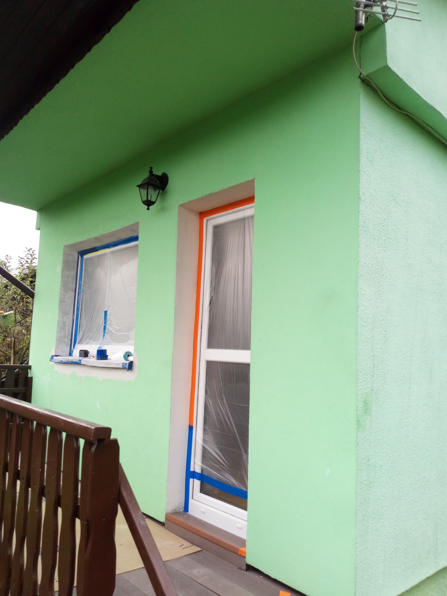 Świeżo pomalowana na zielono elewacja domu z zabezpieczonym oknem i drzwiami folią malarską i taśmą, widoczna lampa zewnętrzna i antena dachowa.