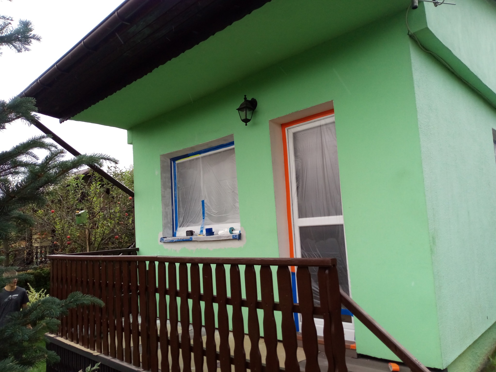Dom jednorodzinny z drewnianą balustradą balkonu, świeżo pomalowany na jasnozielony kolor, okna zabezpieczone folią i taśmą malarską, widoczna lampa zewnętrzna i fragment ogrodu z drzewami owocowymi.