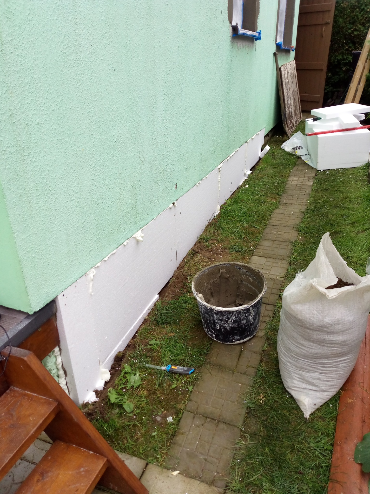 Ocieplanie fundamentu budynku styropianem, widoczny fragment elewacji, wiadro z zaprawą, torba z ziemią oraz ścieżka z kostki brukowej.