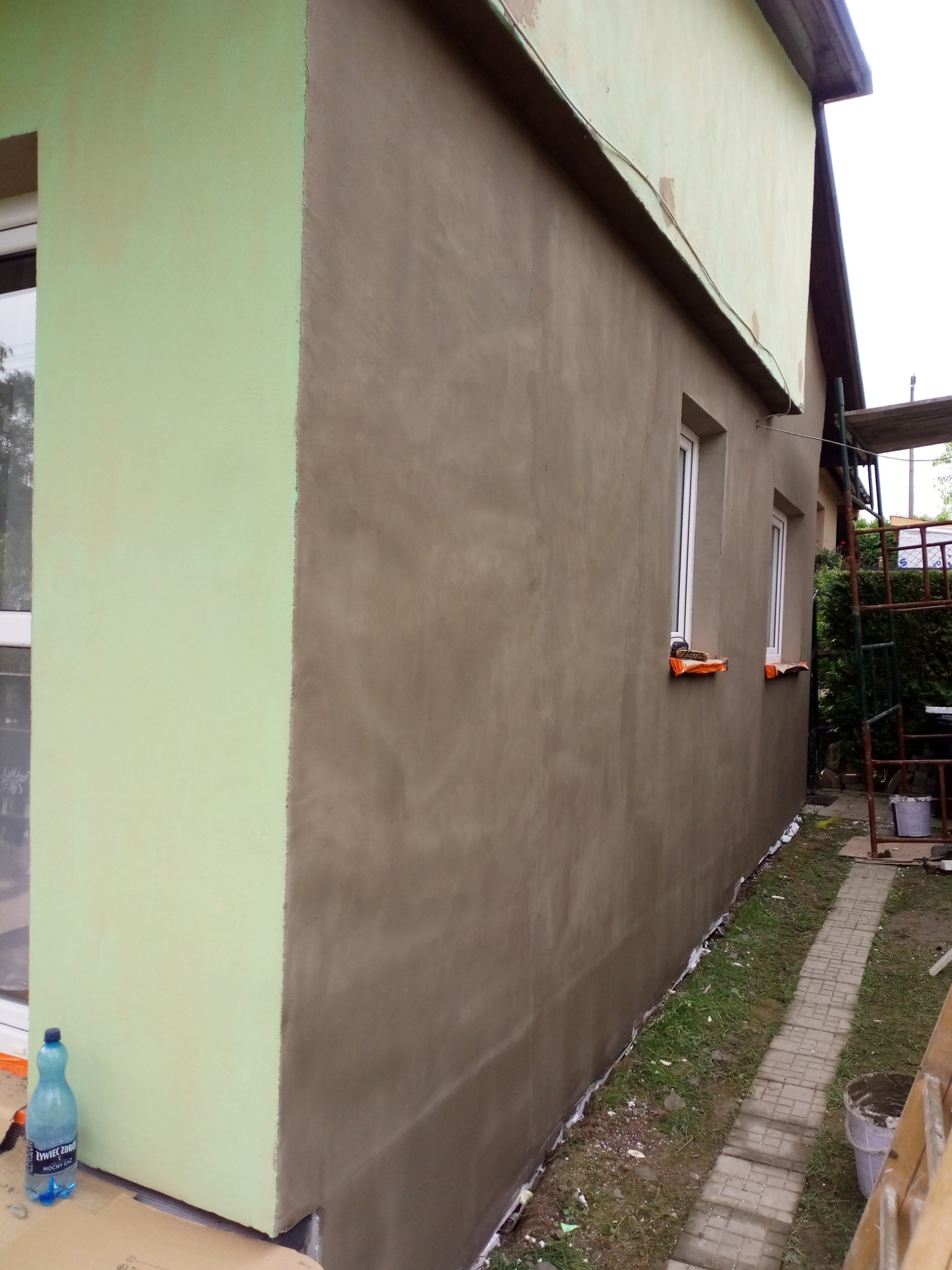 Ściana budynku po nałożeniu nowej warstwy szpachli, z widocznym fragmentem ocieplenia w kolorze pistacjowym, zabezpieczone okna, rusztowanie w tle i butelka wody mineralnej przy ścianie.