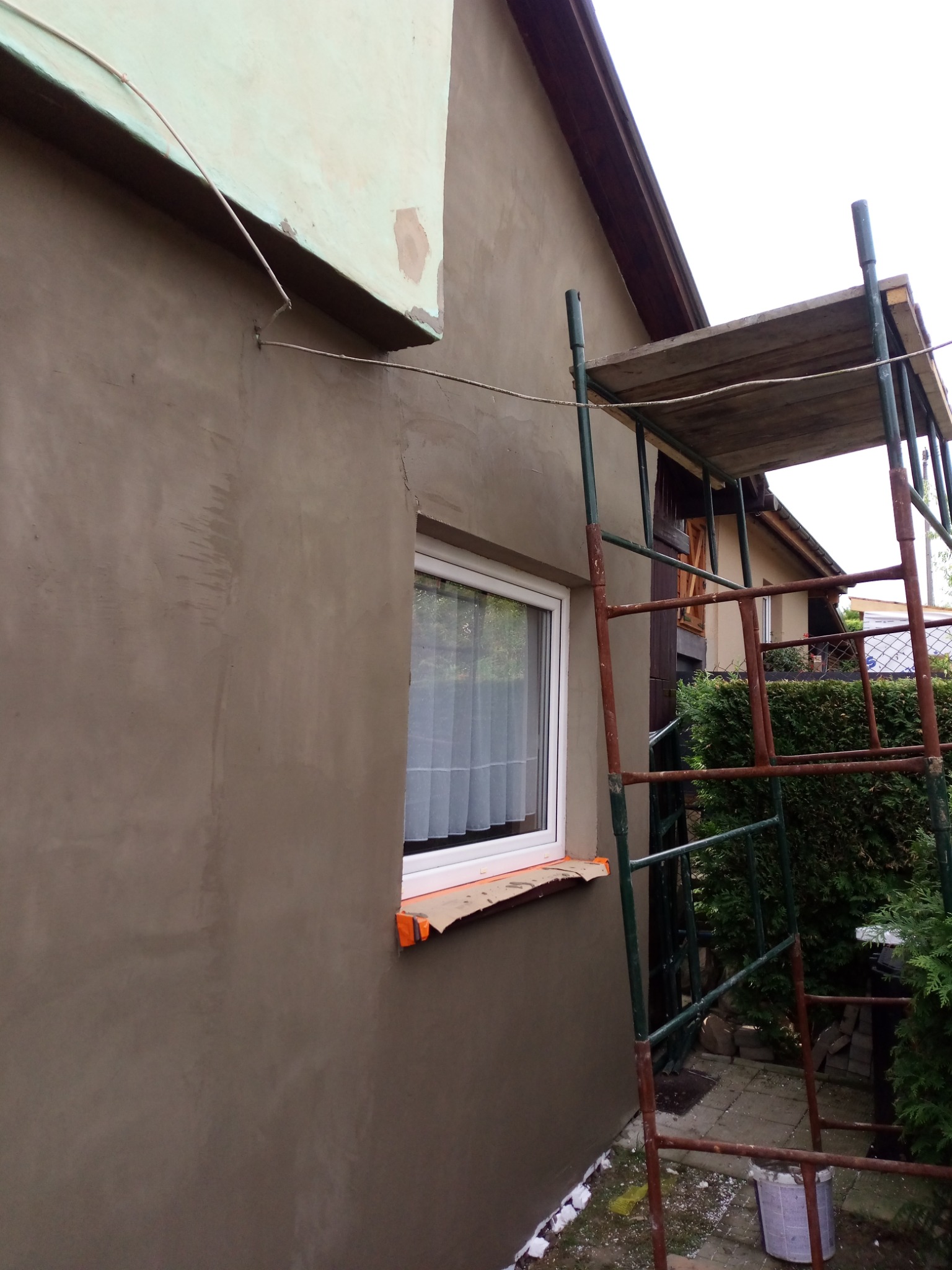 Ściana domu w trakcie nakładania szarego tynku, widoczne rusztowanie z brązowymi elementami i białe okno z zasłonami. Część ściany pokryta zielonkawym materiałem izolacyjnym.