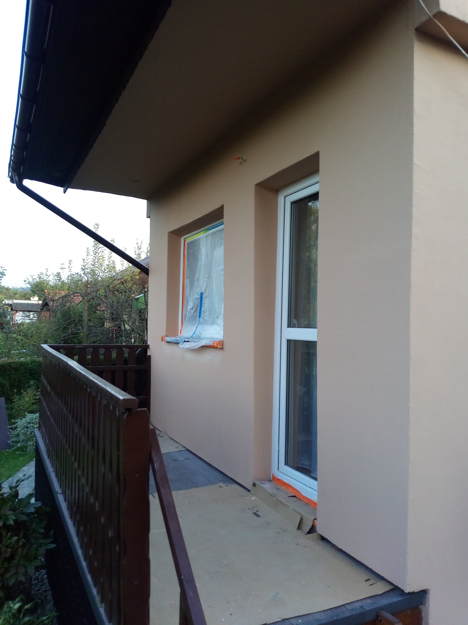 Boczny widok budynku z beżową elewacją, z oknem zabezpieczonym folią malarską, białymi drzwiami balkonowymi i drewnianą balustradą balkonu.