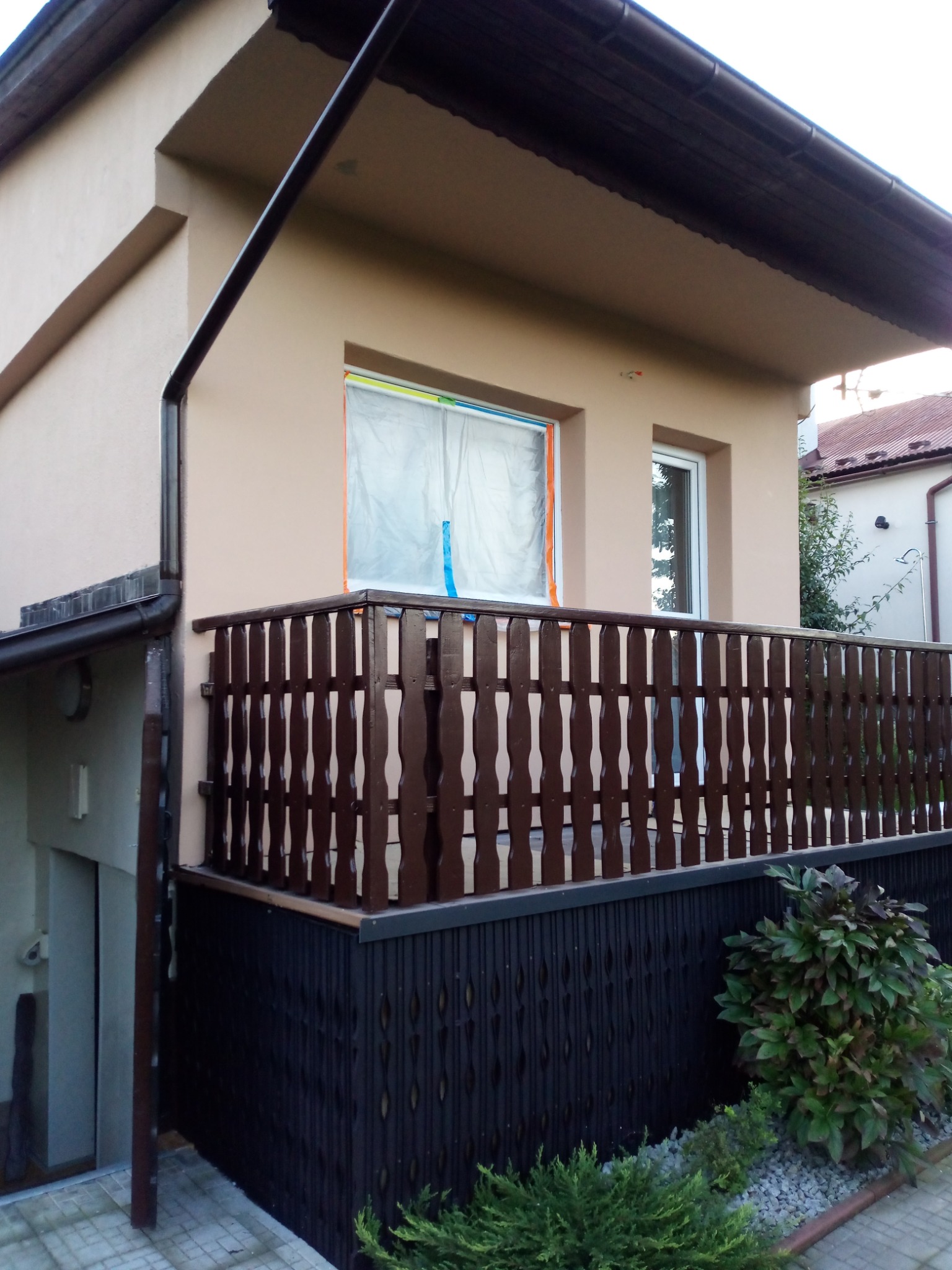 Elewacja budynku z balkonem z brązową balustradą i oknem zabezpieczonym folią malarską, widoczne rynny i fragment dachu.