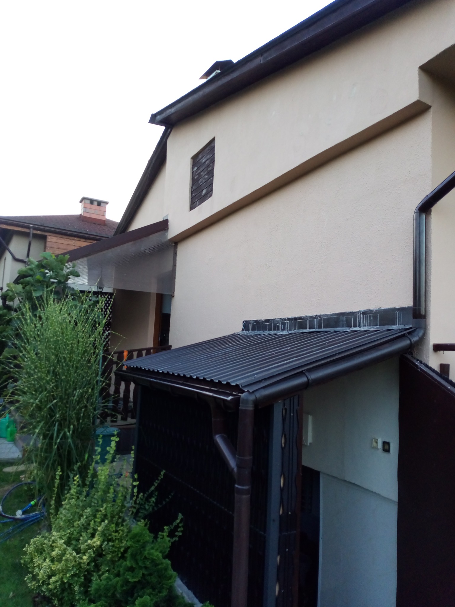 Elewacja domu z widoczną zabudową nad wejściem, rynny spustowe i fragment ogrodu z roślinnością