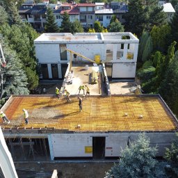 New Structure - Wysoki kąt widzenia na budowę domu z betonu komórkowego; widoczny strop z ułożonym zbrojeniem, pracownicy w kamizelkach odblaskowych, pompa do betonu w akcji i częściowo wykończony budynek obok...