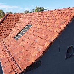 New Structure - Nowy dach z czerwonej dachówki karpiówki na szarym budynku z oknem w kształcie łuku, widoczna łatka w miejscu okna dachowego.