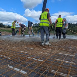 New Structure - Wylewanie betonu na przygotowane zbrojenie fundamentów przez grupę pracowników w odblaskowych kamizelkach na tle błękitnego nieba z chmurami.
