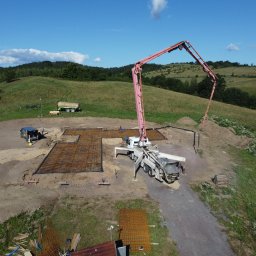 New Structure - Widok z drona na plac budowy z przygotowanym zbrojeniem fundamentów i pompą do betonu na tle pagórkowatego krajobrazu w słoneczny dzień.
