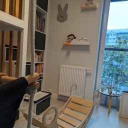 New Structure - Jasny pokój dziecięcy z drewnianą huśtawką w kształcie łódki, zegarem w kształcie królika i półkami w kształcie chmur z zabawkami, widok na blok przez okno.