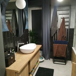 New Structure - Nowoczesna łazienka z owalną umywalką nablatową, drewnianą szafką pod umywalką, czarnymi płytkami ściennymi, lustrem w dekoracyjnej ramie i grzejnikiem drabinkowym z ręcznikami.