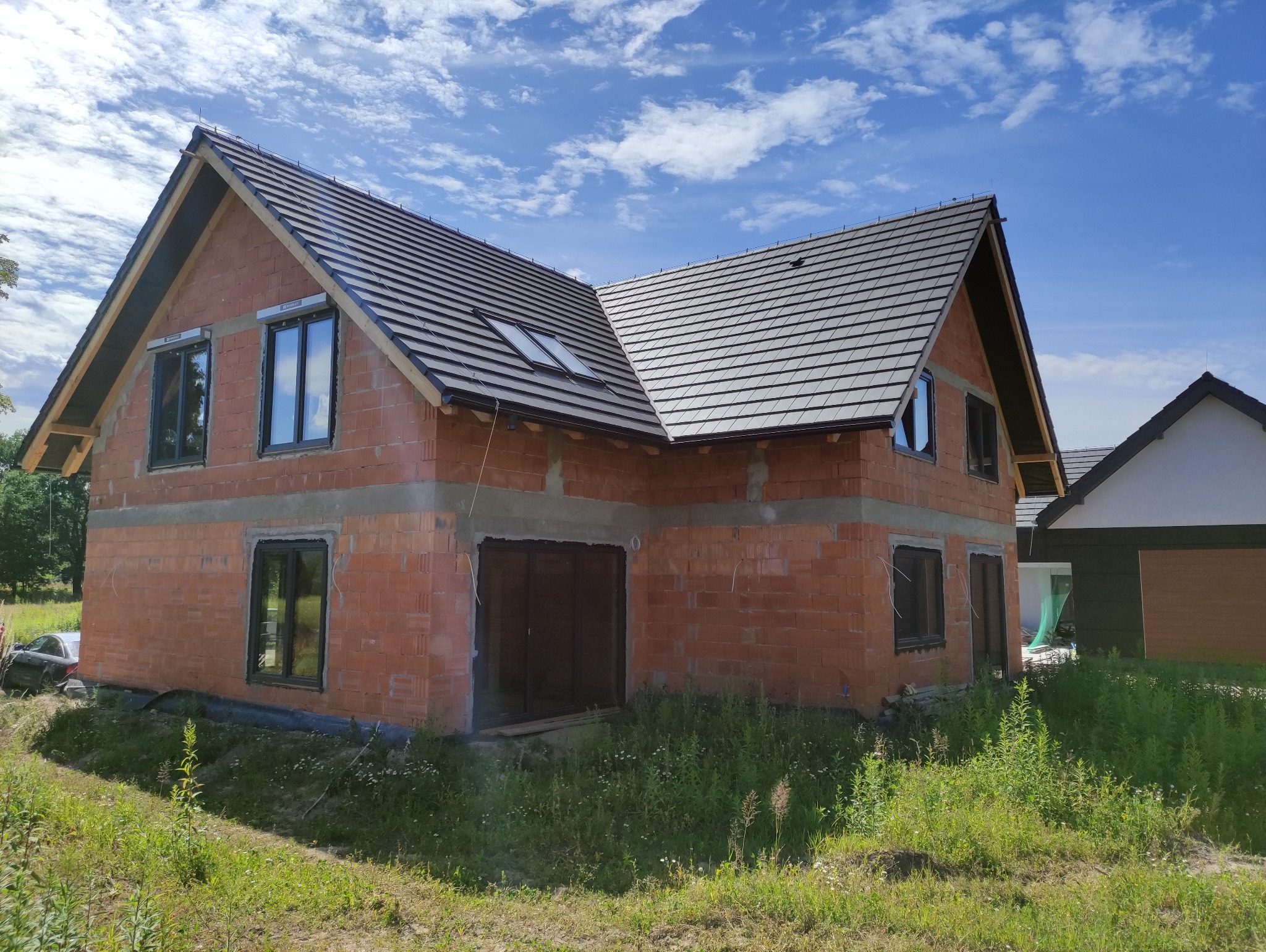 Surowy, dwukondygnacyjny dom murowany z czerwonej cegły w trakcie budowy, z zamontowanymi oknami i ciemną dachówką, widoczne nieotynkowane ściany i dachowe okna, w tle błękitne niebo i zarośnięty...