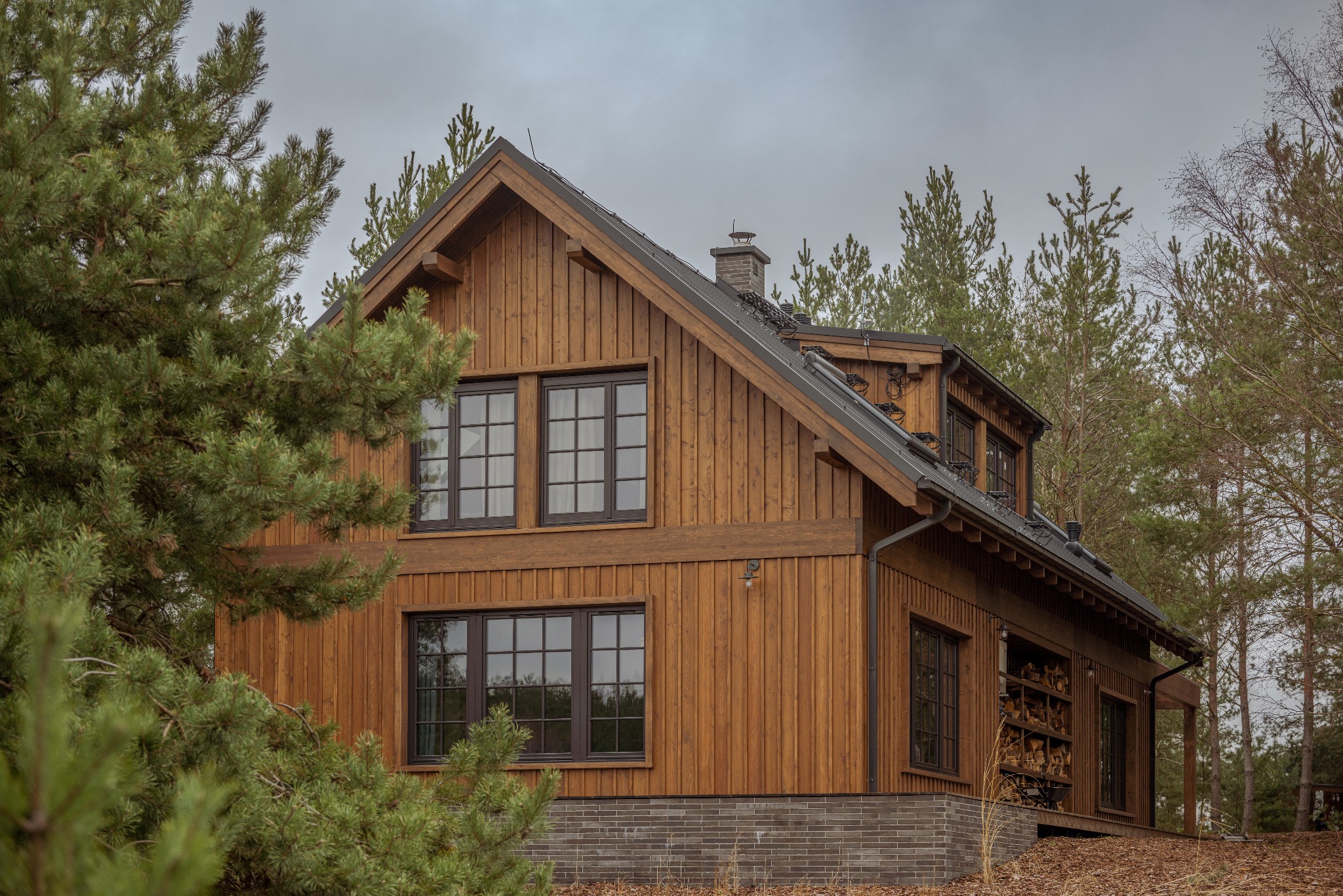 Dwukondygnacyjny dom z drewnianą elewacją, z czarnymi ramami okiennymi i szarym dachem, częściowo zasłonięty przez sosnę, z widocznym składem drewna pod okapem dachu.