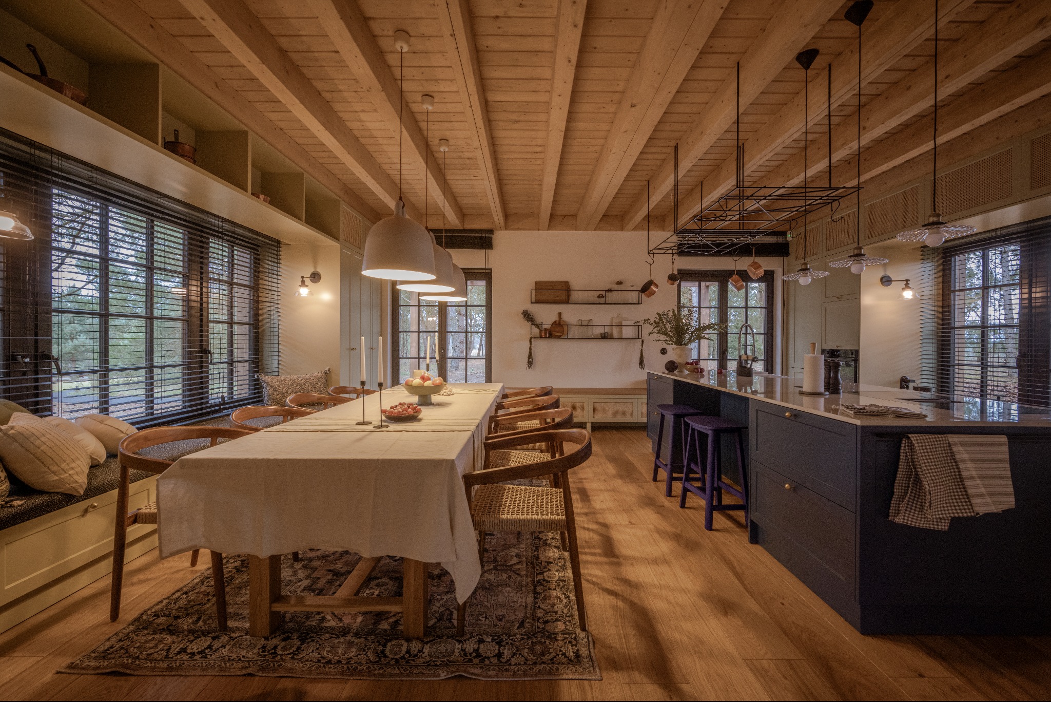 Jasna kuchnia z drewnianym stropem, dużym oknem z żaluzjami, wyspą kuchenną z fioletowymi stołkami barowymi i stołem jadalnym nakrytym białym obrusem.