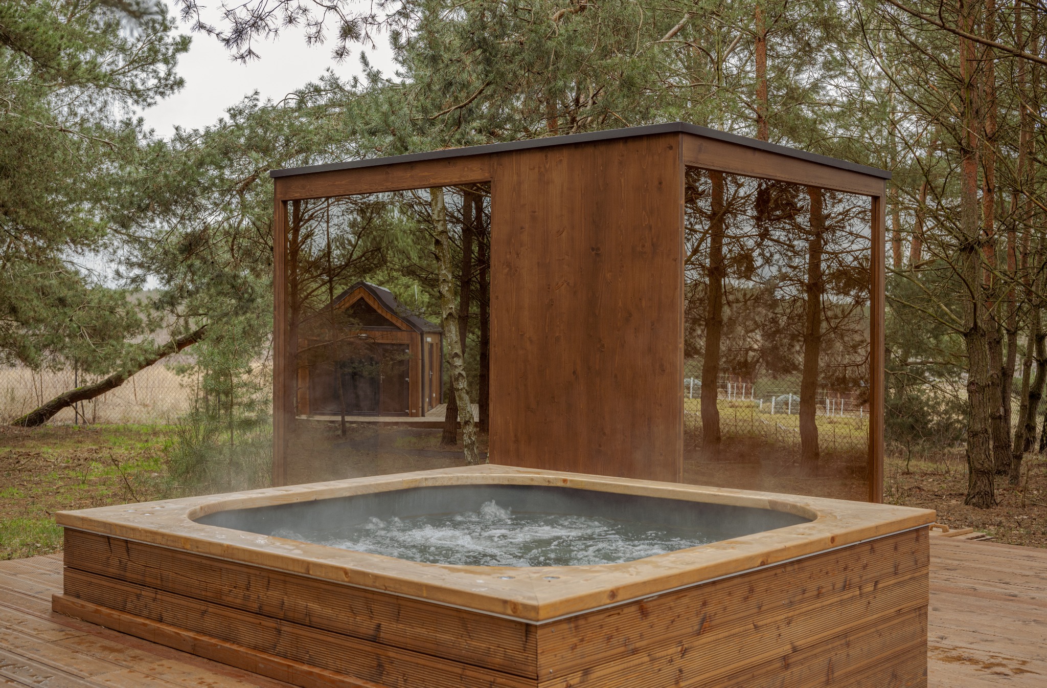 Drewniana balia z wodą na drewnianym tarasie, otoczona lasem i minimalistyczną konstrukcją z lustrzanymi ścianami, w których odbija się mały drewniany domek.