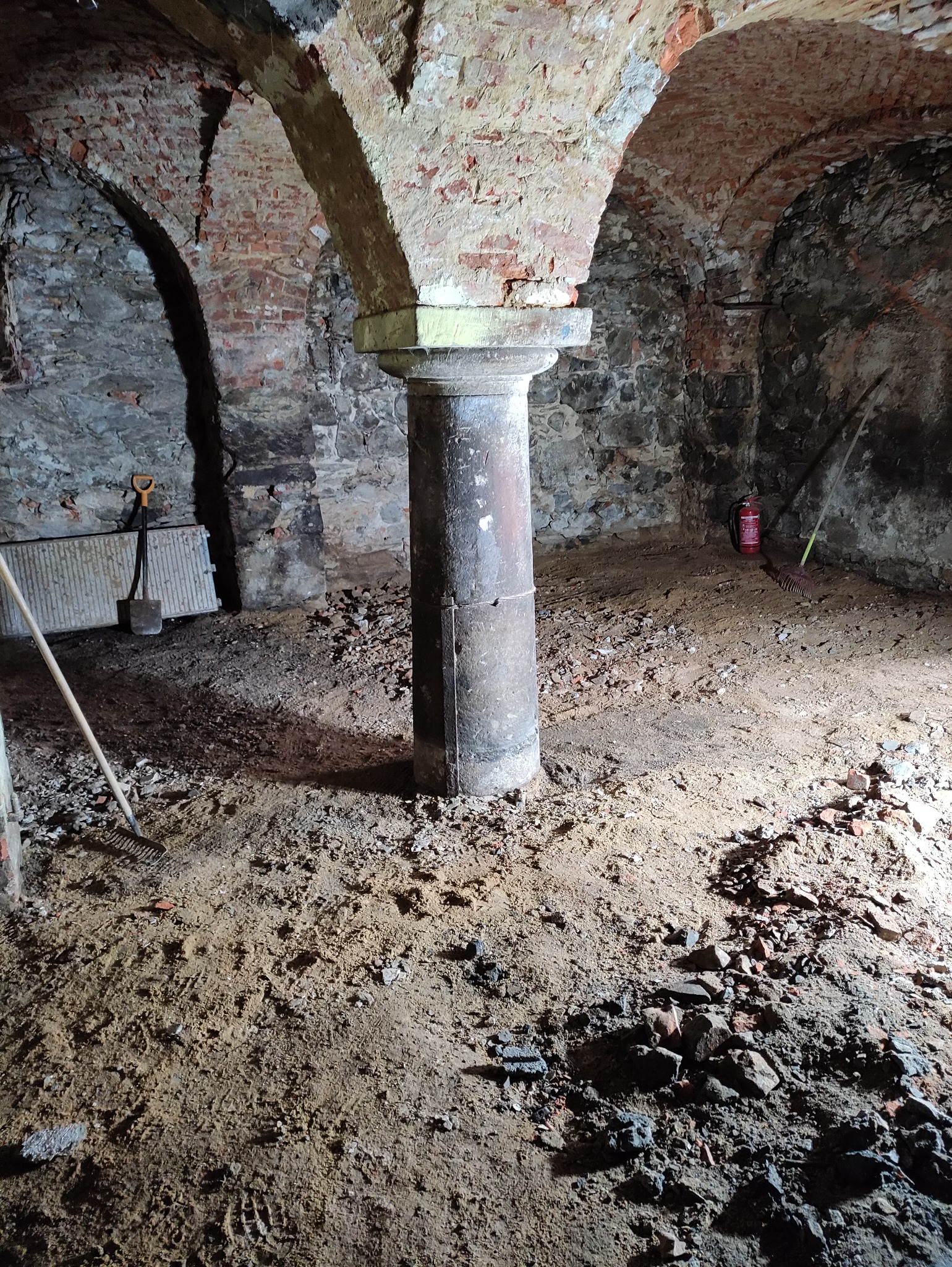 Wnętrze piwnicy z kolumną wspierającą sklepienie łukowe z cegły, widoczne narzędzia budowlane: łopata, grabie, oraz gaśnica w tle, podłoże pokryte ziemią i gruzem.
