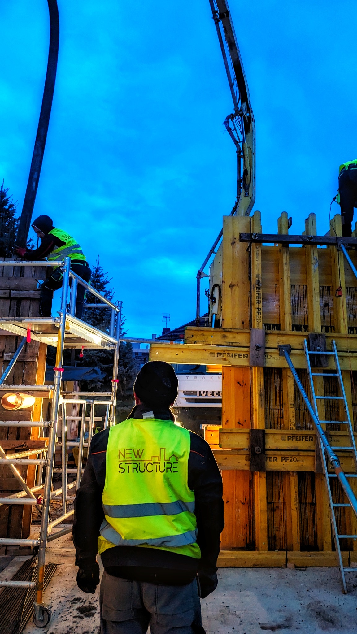 Pracownicy w odblaskowych kamizelkach przy szalunku fundamentów, w tle pompa do betonu i ciężarówka Iveco, wieczorne niebieskie niebo.