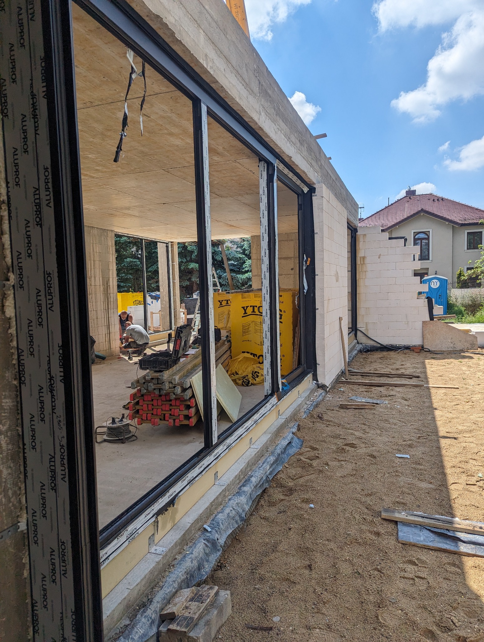 Budowa domu w stanie surowym otwartym z zamontowanymi oknami panoramicznymi Aluplast, widoczne bloczki Ytong, elementy zbrojeniowe i pracujący robotnik wewnątrz budynku.