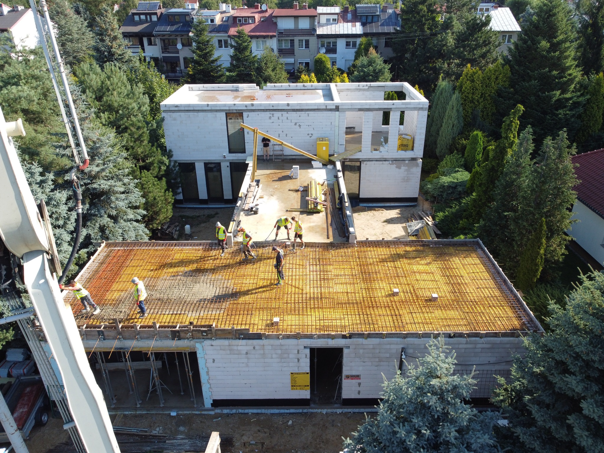 Wysoki kąt widzenia na budowę domu z betonu komórkowego; widoczny strop z ułożonym zbrojeniem, pracownicy w kamizelkach odblaskowych, pompa do betonu w akcji i częściowo wykończony budynek obok...