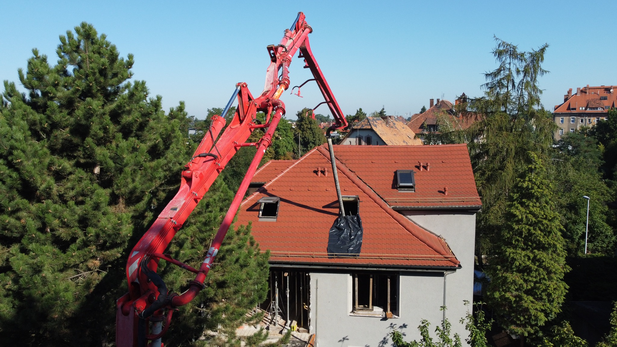 Czerwona pompa do betonu podaje mieszankę przez otwór w dachu remontowanego domu z czerwonej dachówki, widok z góry.