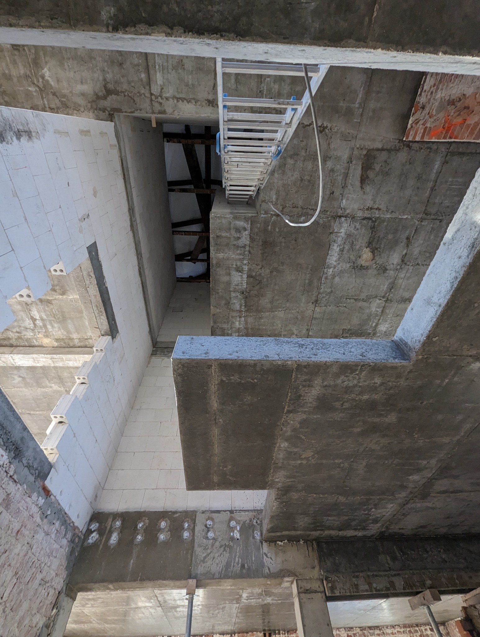 Wnętrze surowego budynku w trakcie budowy, widoczne betonowe stropy, ściany z białego bloczka, drabina aluminiowa i fragment ściany z czerwonej cegły z pomarańczowymi oznaczeniami.