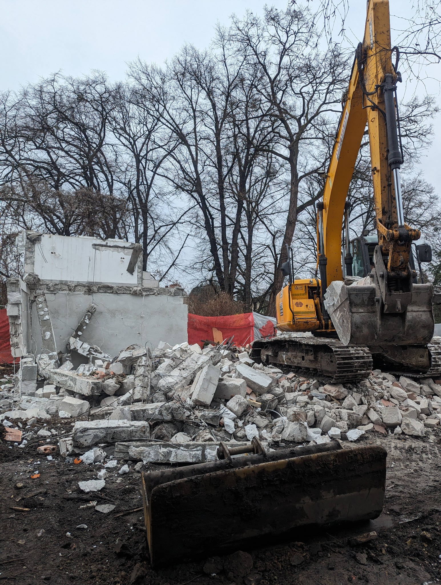 Żółta koparka gąsienicowa na tle zrujnowanego budynku z cegieł i betonu, widoczne fragmenty ścian i gruzowisko, w tle nagie drzewa i pochmurne niebo.