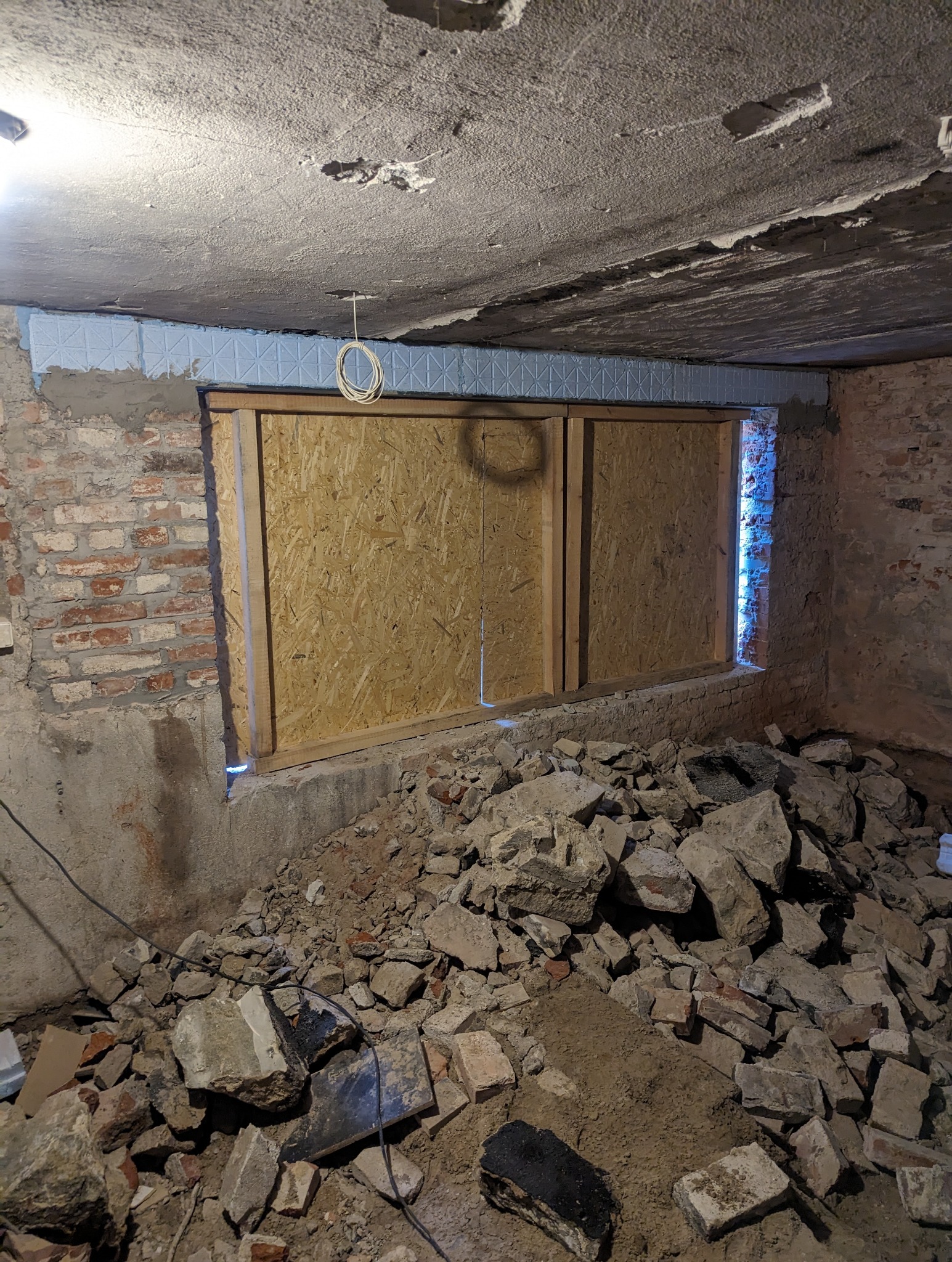 Piwnica w trakcie remontu, zabetonowane okno z deskami OSB, gruz na podłodze, widoczne ślady po skuciu tynku, izolacja termiczna na suficie.