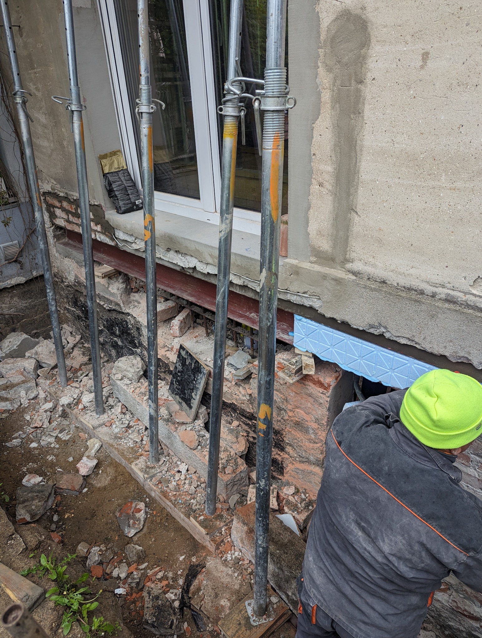 Prace przy wzmocnieniu fundamentów domu z widocznymi podporami stalowymi, wykopem odsłaniającym fragment fundamentu z ociepleniem z niebieskiego styroduru oraz pracownikiem w neonowej czapce.