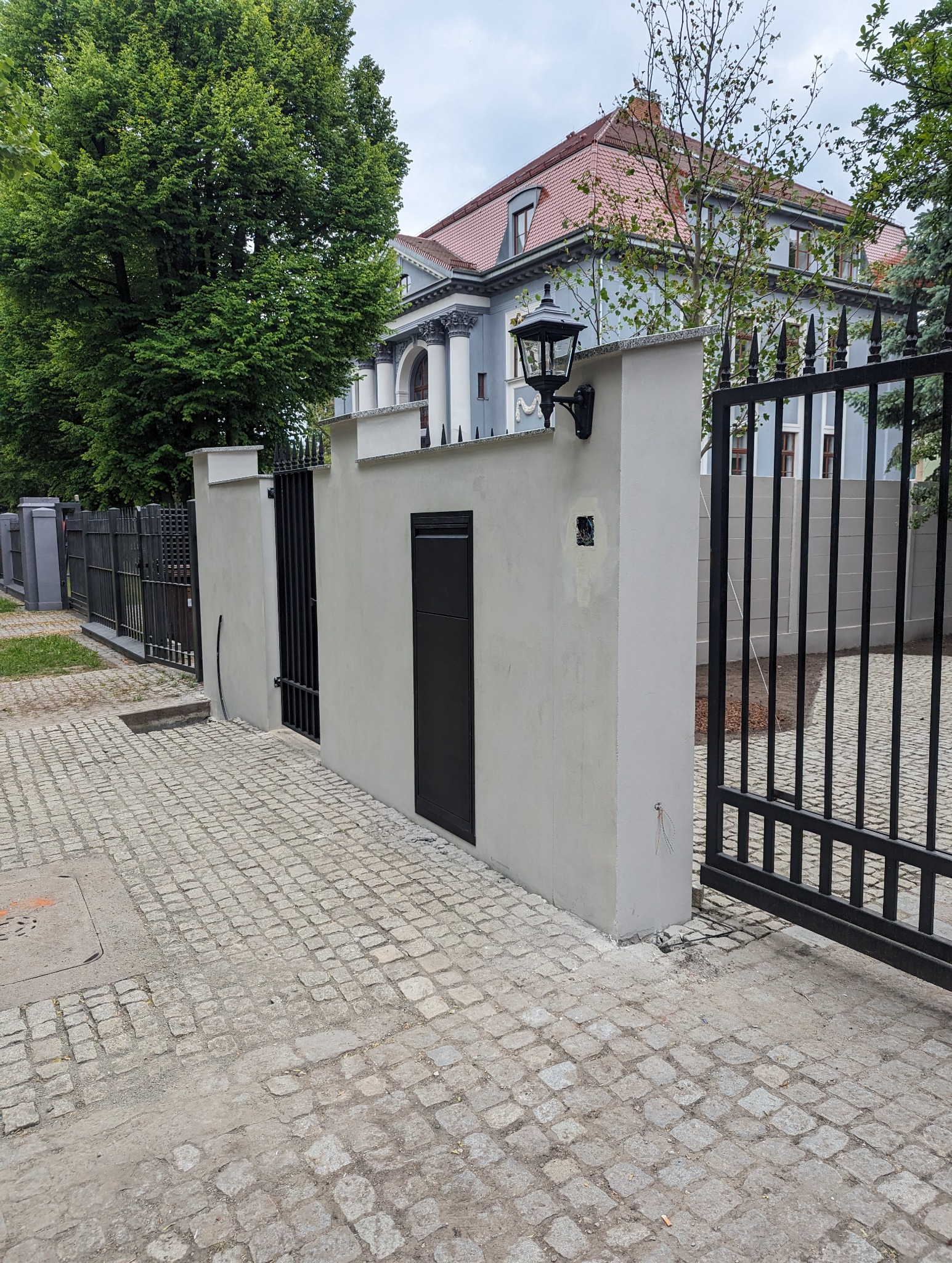 Nowoczesne ogrodzenie z szarego betonu z czarną skrzynką na listy i lampą, brama z czarnego metalu, kostka brukowa na podjeździe, w tle duży dom z czerwonym dachem i kolumnami.