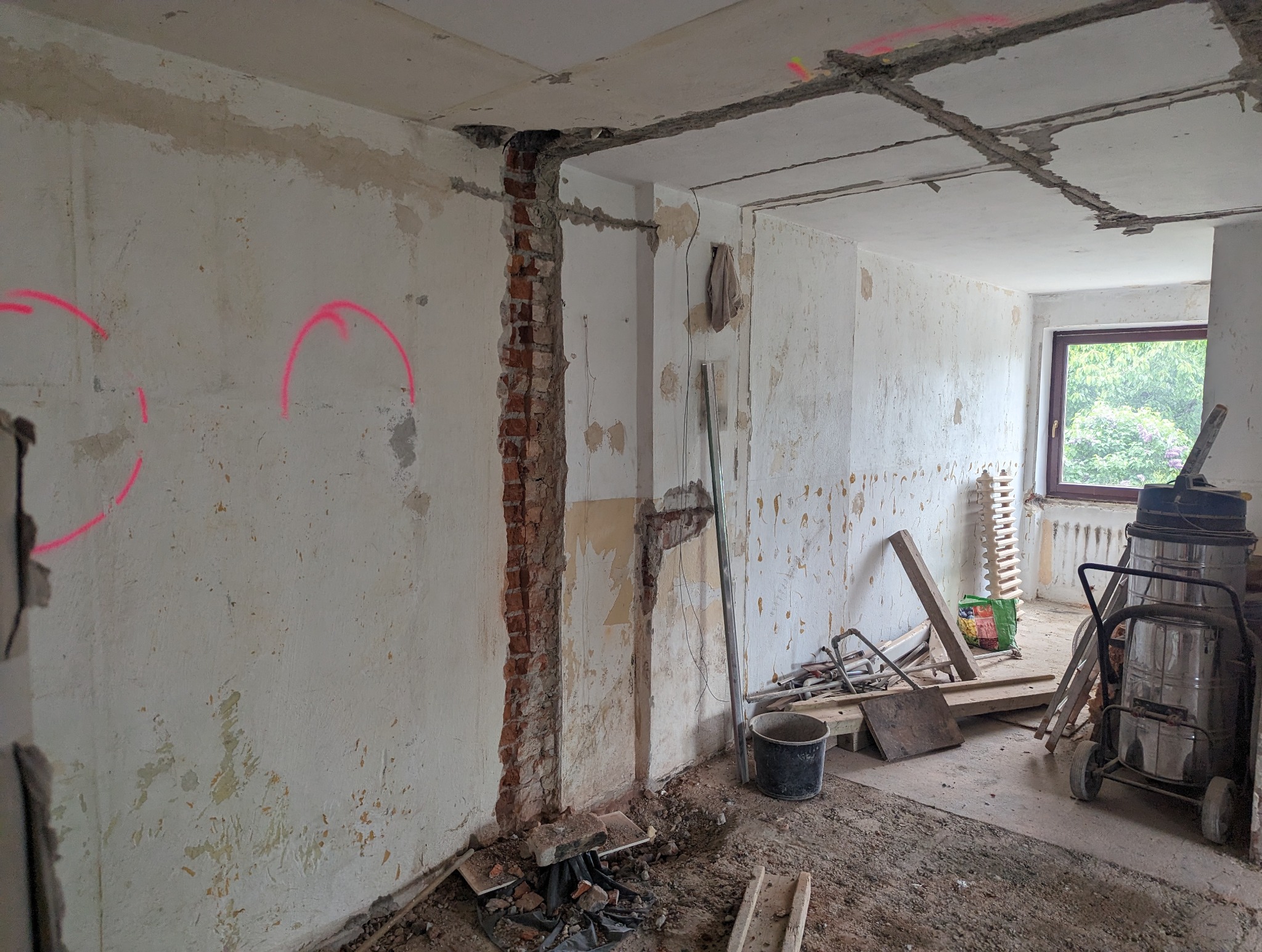 Wnętrze w trakcie remontu: odsłonięta cegła, ściany ze śladami farby, oznaczenia różową farbą, gruz na podłodze, widoczne narzędzia i odkurzacz przemysłowy przy oknie.