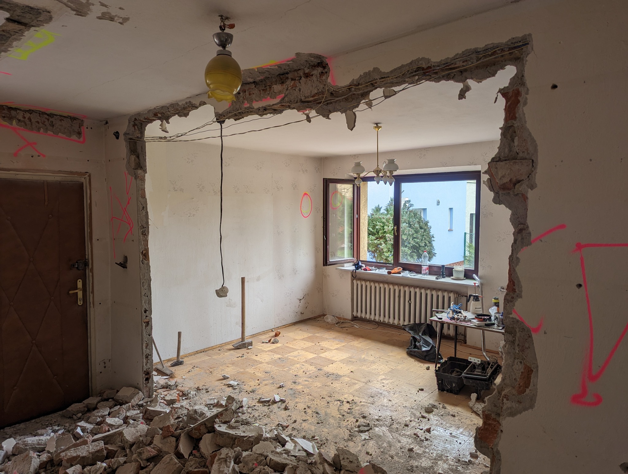 Demontaż ściany w mieszkaniu, widoczne gruzowisko, odsłonięte przewody elektryczne, ślady po wyburzeniach zaznaczone różowym markerem, narzędzia na podłodze, grzejnik pod oknem.