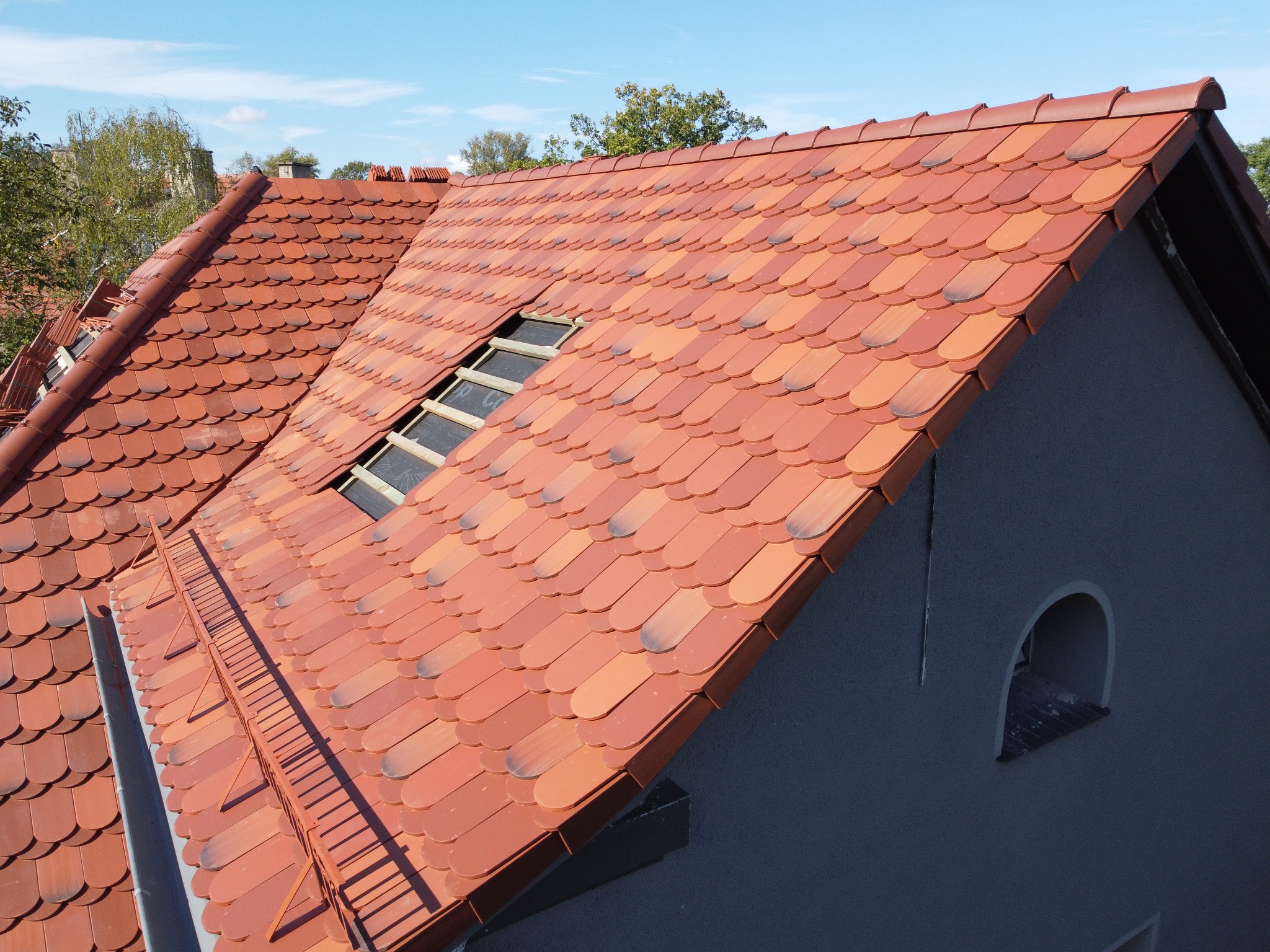 Nowy dach z czerwonej dachówki karpiówki na szarym budynku z oknem w kształcie łuku, widoczna łatka w miejscu okna dachowego.