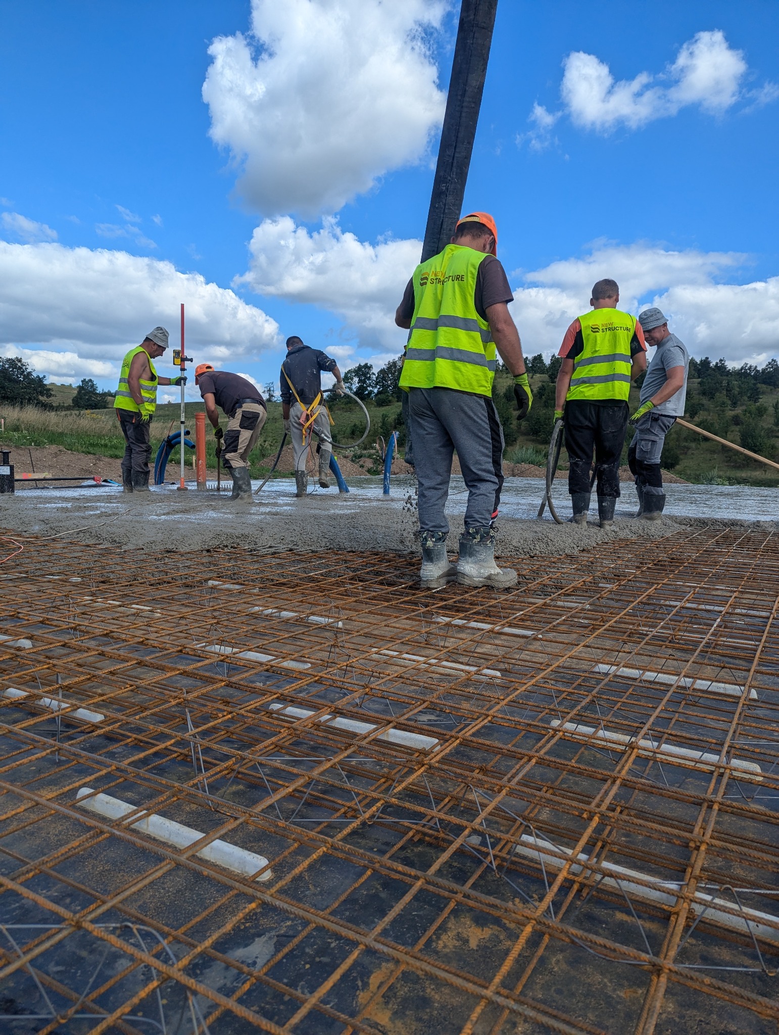 Wylewanie betonu na przygotowane zbrojenie fundamentów przez grupę pracowników w odblaskowych kamizelkach na tle błękitnego nieba z chmurami.