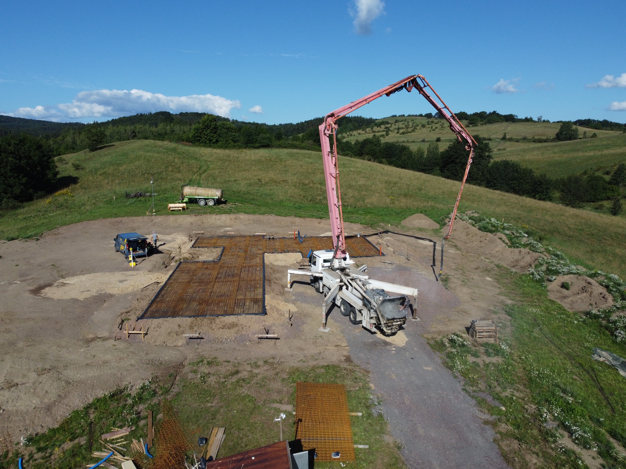 Widok z drona na plac budowy z przygotowanym zbrojeniem fundamentów i pompą do betonu na tle pagórkowatego krajobrazu w słoneczny dzień.
