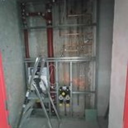 usługi ogólnobudowlane Jakub Sura - Wnętrze w trakcie montażu instalacji wodnej: widoczne rury miedziane, metalowa konstrukcja wsporcza, rozdzielacze z zaworami i drabina aluminiowa. Ściany surowe, betonowe.